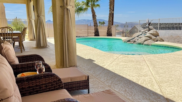 Un espace piscine aménagé avec des meubles et des coussins en osier, offrant une vue sur les montagnes, et entouré d’une clôture.