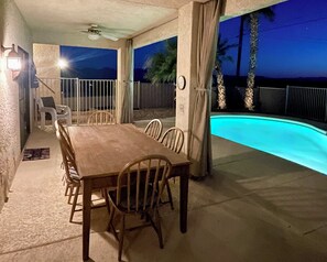 Ein überdachter Außenbereich zum Essen mit Pool, ausgestattet mit einem Holztisch und -stühlen, einem Deckenventilator und Blick auf den Pool bei Nacht.