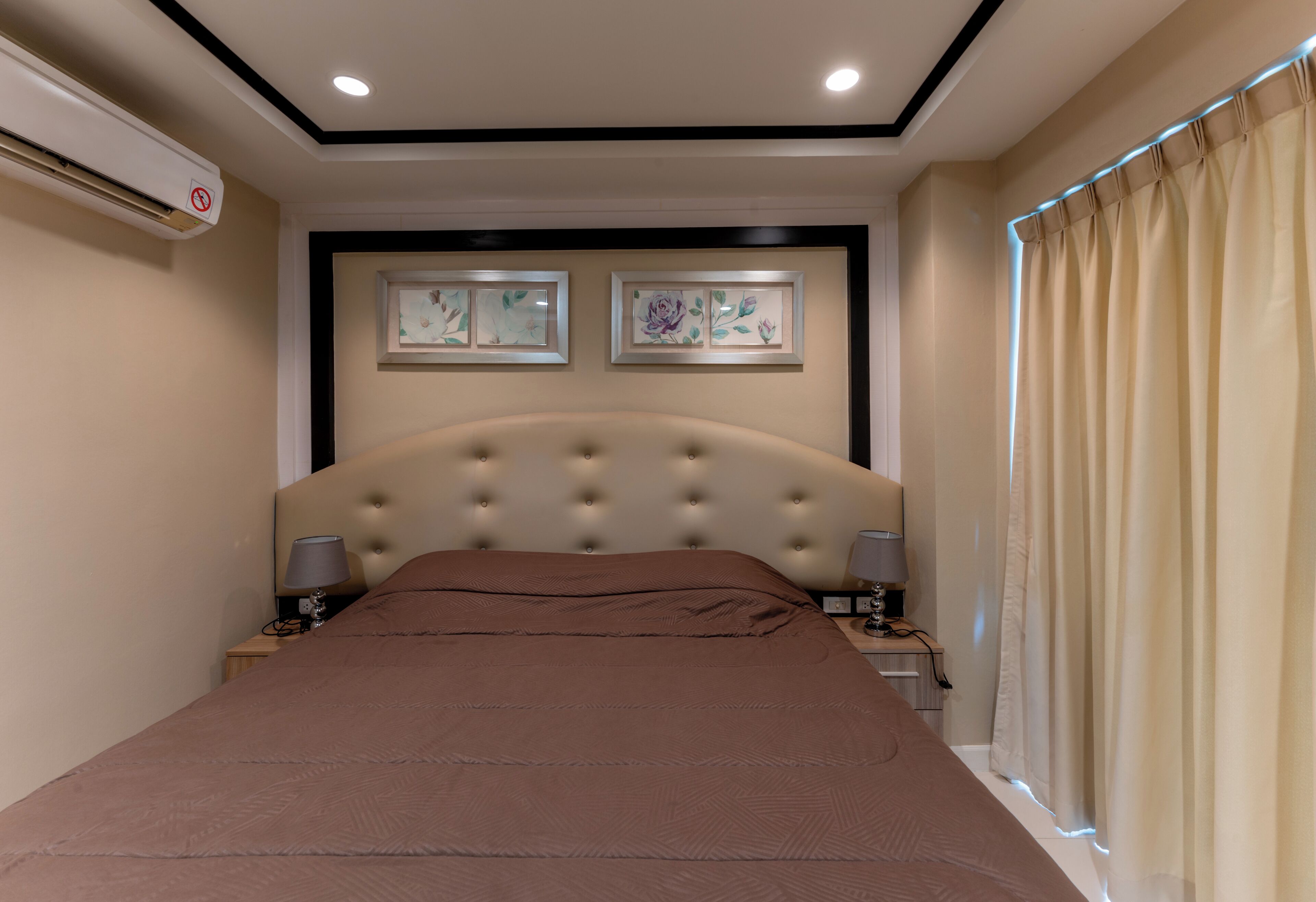 A neatly arranged bedroom with a large bed, two bedside tables with lamps, a wall-mounted air conditioner, and framed artwork above the bed.