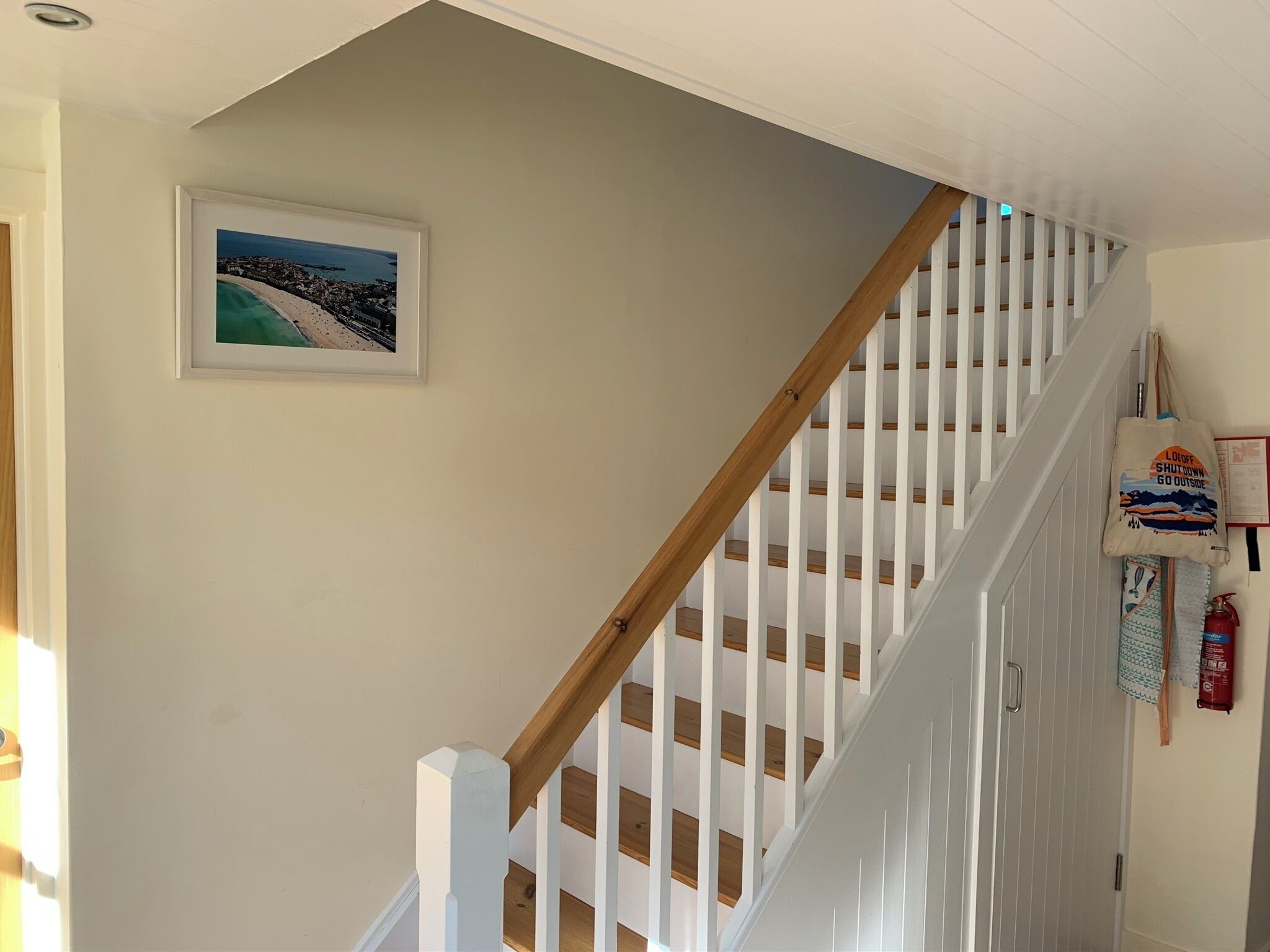 Un escalier avec des rampes blanches et des marches en bois, un tableau encadré accroché au mur et un extincteur sur la droite.
