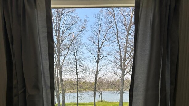 A window with black curtains, a view of a grassy area, trees, and a body of water.