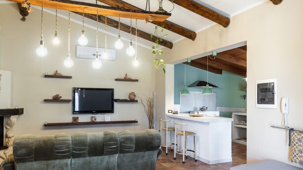 A cozy living room with a sofa, a TV, and hanging lights. There's a kitchen area with a bar and stools. - Villa Scrusciu Ri Mari with private pool, Noto (Sicilia)