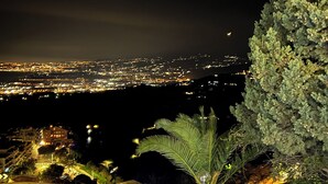 Une vue nocturne depuis un balcon, avec un paysage urbain en arrière-plan, un palmier et un escalier doté de rampes.