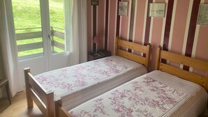 A room with two wooden beds, a window with curtains, a small bedside table with a lamp, and a view of a green landscape. - Mountain chalet   (Mont-Dore)