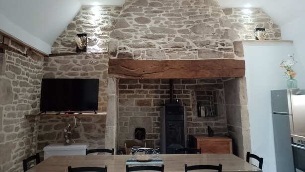Un comedor con pared de piedra, una mesa de madera, sillas, un televisor de pantalla plana y una estufa de leña empotrada.