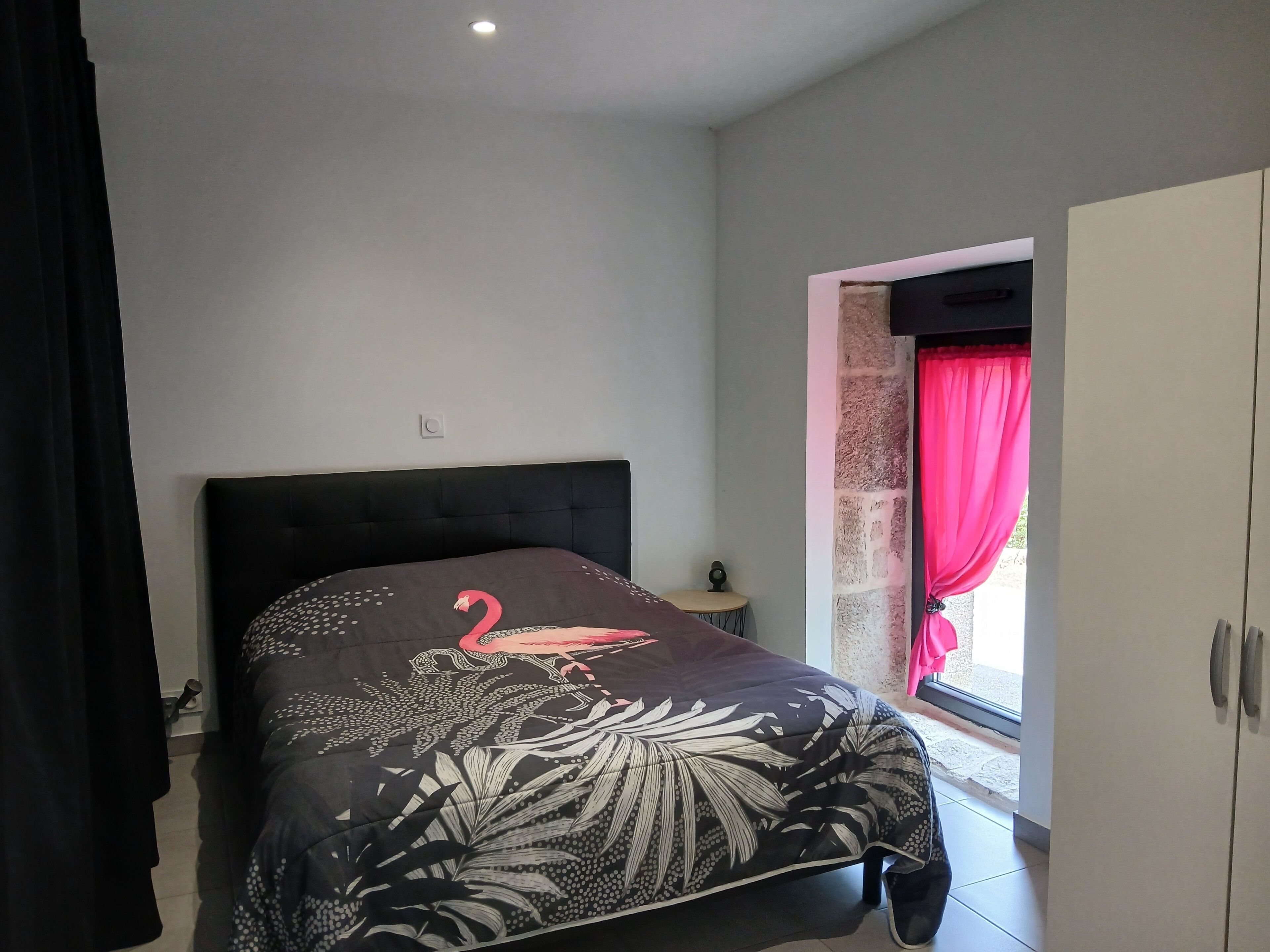Un dormitorio con cama, mesita de noche, armario y ventana con cortinas.