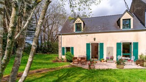 A two-story house with a front porch, green shutters, and a small garden area with potted plants and a table. - Le Relais - Maison avec grand jardin (Bannalec)