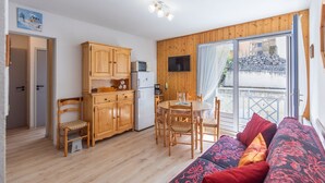 En kompakt opholdsstue med trægulv, spiseplads med bord og stole, et tekøkken med mikroovn og køleskab, samt en sofa med mønstrede puder.