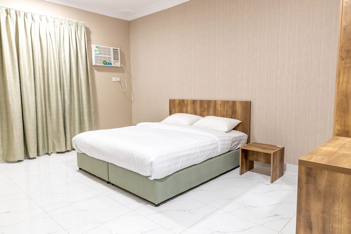 A bedroom with a bed, a nightstand, a wooden headboard, a wall-mounted air conditioner, and a curtain.