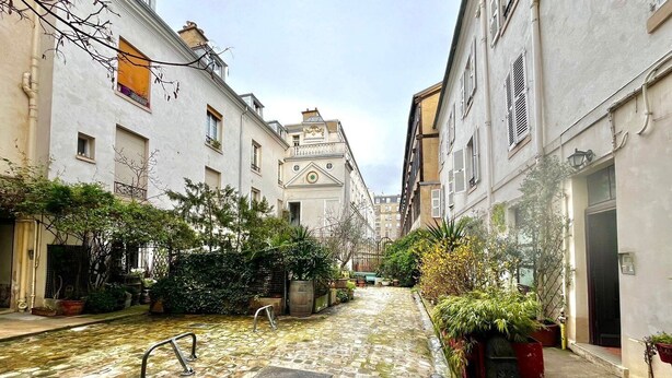 Une ruelle étroite pavée, bordée d’immeubles dotés de balcons et de plantes en pot.