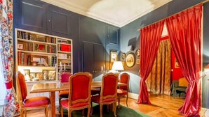 Un coin repas avec une table en bois et des chaises à revêtement rouge, une bibliothèque remplie de livres et de photos encadrées, et un rideau rouge à la fenêtre.