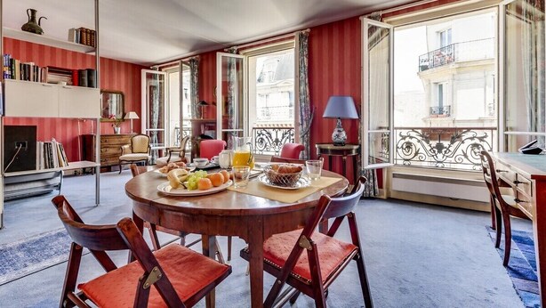 Un coin repas avec une table ronde dressée pour le petit-déjeuner, entourée de chaises. On y trouve également des étagères et une vue sur le balcon extérieur.