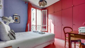 Une chambre à coucher avec un lit, une table en bois et une chaise, une armoire rouge et une fenêtre avec des rideaux.