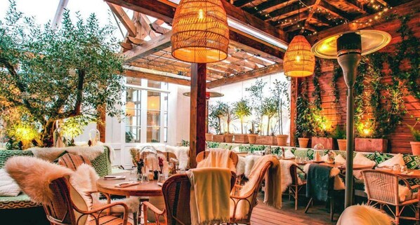 Un restaurante al aire libre con mobiliario de mimbre, lámparas colgantes y plantas en macetas.