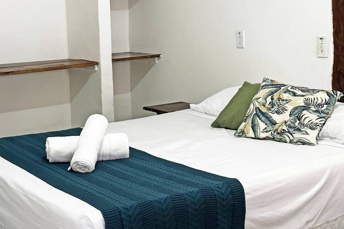 A neatly made bed with a teal blanket, a green pillow, and rolled white towels. There are wooden shelves on the wall and a nightstand beside the bed.
