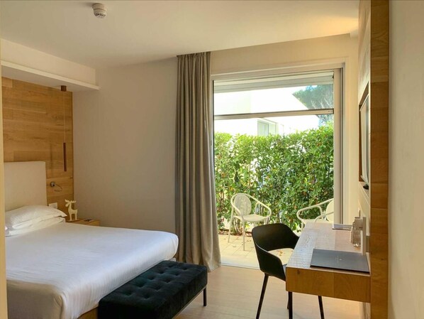 Un dormitorio con cama, escritorio y silla. Hay una ventana con cortinas, y un balcón con sillas y plantas afuera.