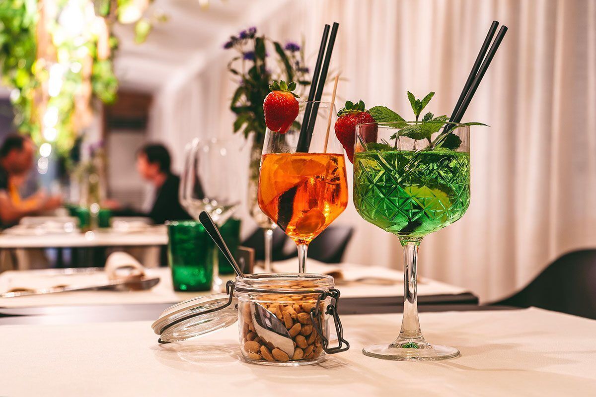 Two cocktails on a table with a jar of nuts and a glass of green liquid with mint leaves.