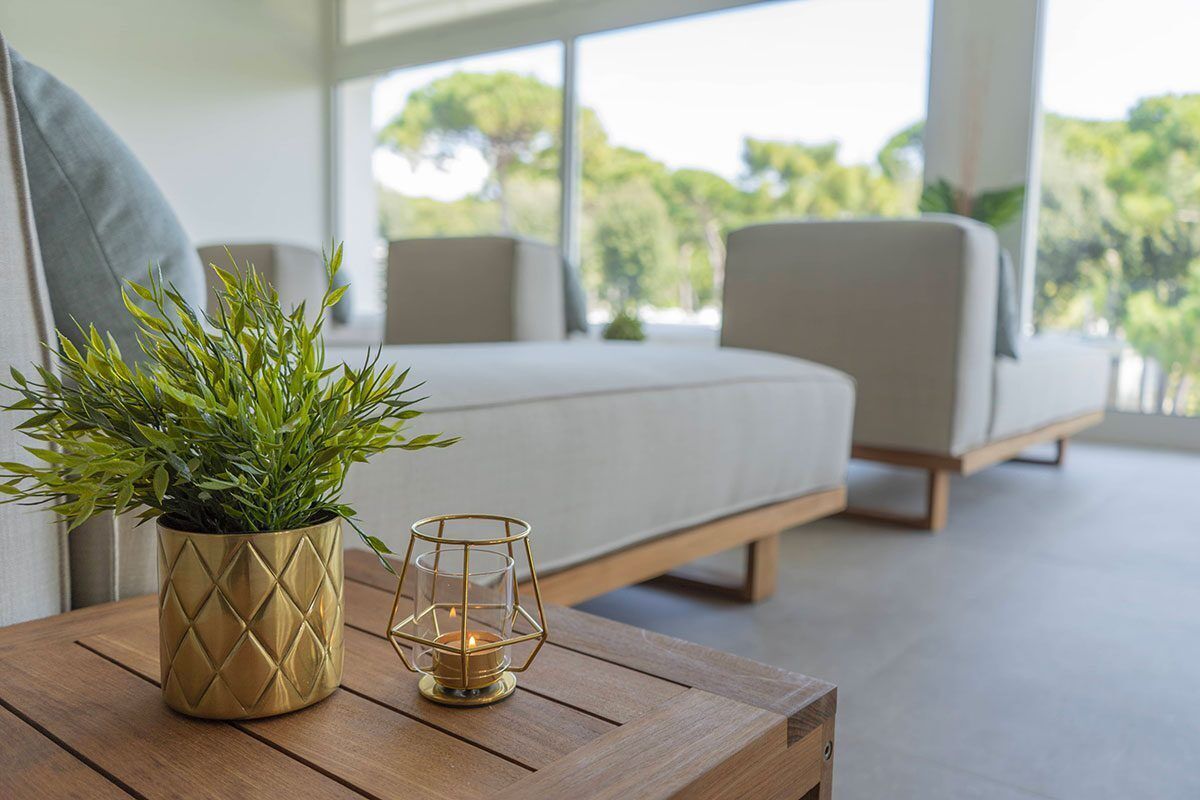 A modern room with a bed, a small table with a plant and candle, and a view of trees through large windows.