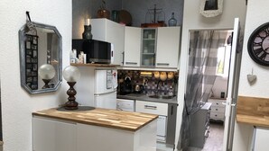 A compact kitchen with white cabinets, a wooden countertop, and a small island. There is a refrigerator, a microwave, and a sink. A clock is visible on the wall. - Joli Petit Mazet Avec Terrasse en Pleine Nature (COGOLIN)