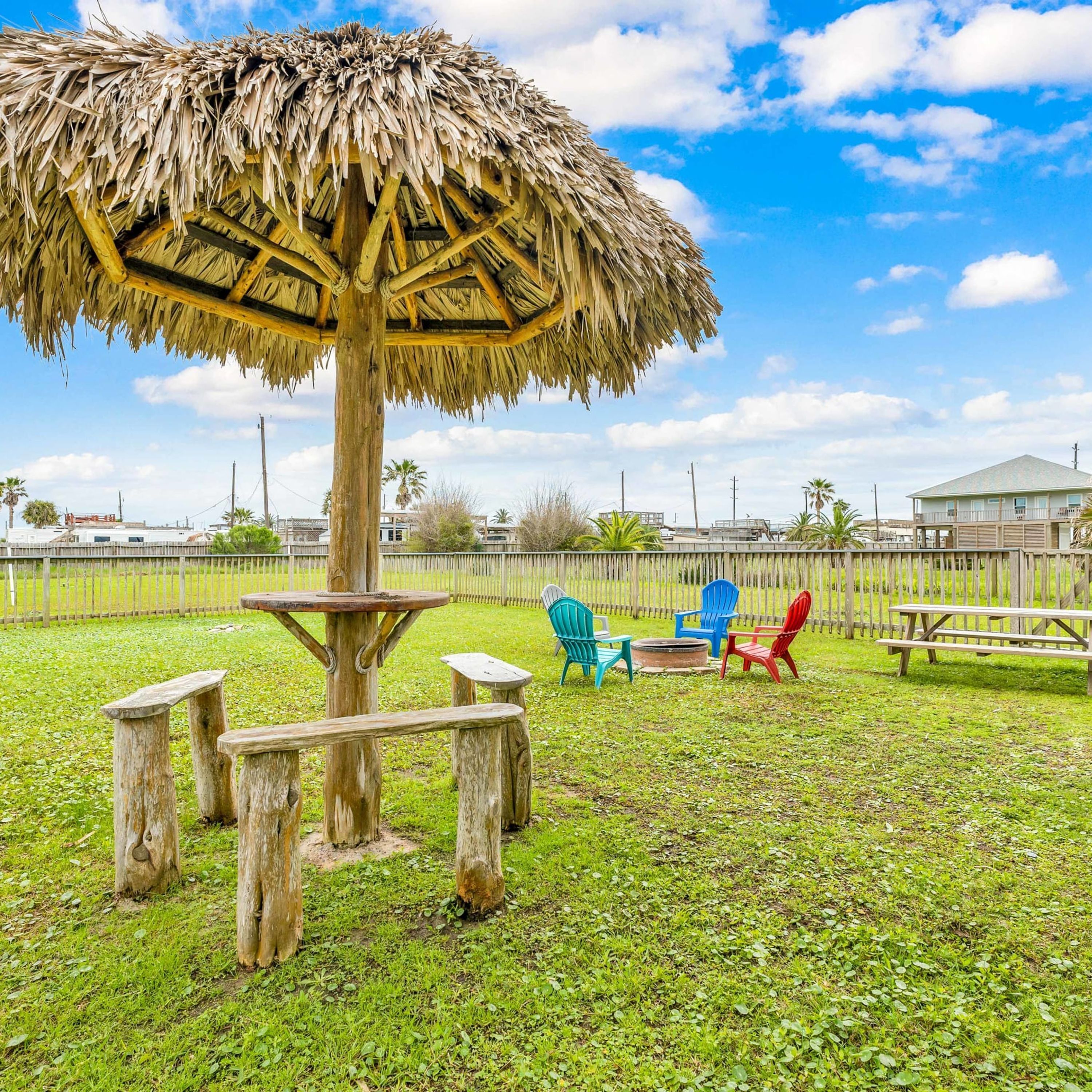 Eine strohgedeckte Sonnenschirmkonstruktion in einer Wiese mit einem Picknicktisch und Stühlen.