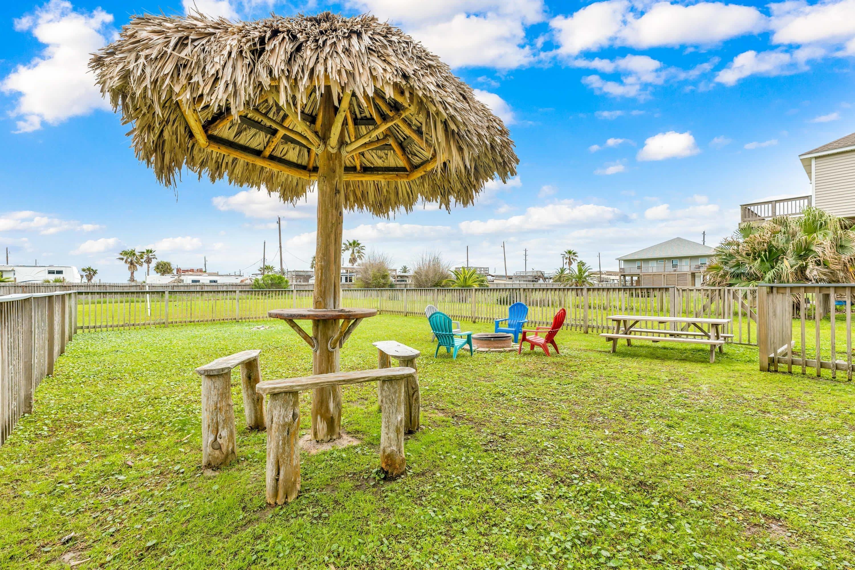 Ein Garten mit einem Reetdachschirm, Holzbänken, Picknicktischen und bunten Stühlen.