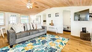 A spacious living room with a gray sofa, a colorful floral rug, wooden ceiling beams, and a ceiling fan. - Cute Beach Bungalow Walk to the Beach (Galveston)