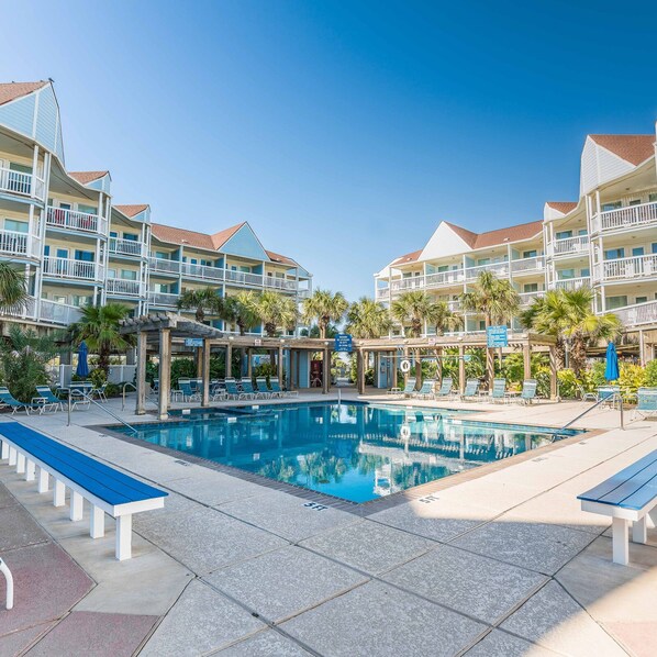 A hotel with a swimming pool, multiple balconies, and palm trees. - Great Pools and Easy Access to Ocean - Get Beach Dazed (Galveston)