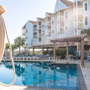 A swimming pool with lounge chairs and a covered area, surrounded by a multi-story building with balconies. - Great Pools and Easy Access to Ocean - Get Beach Dazed (Galveston)