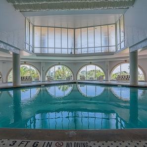 An indoor swimming pool with clear water, surrounded by lounge chairs and a high ceiling with large windows.