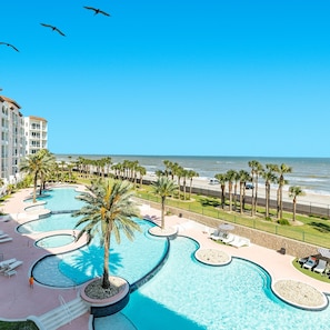 A resort with a large, winding pool surrounded by palm trees and lounge chairs. The pool is adjacent to a beach with a fence separating it from the ocean.