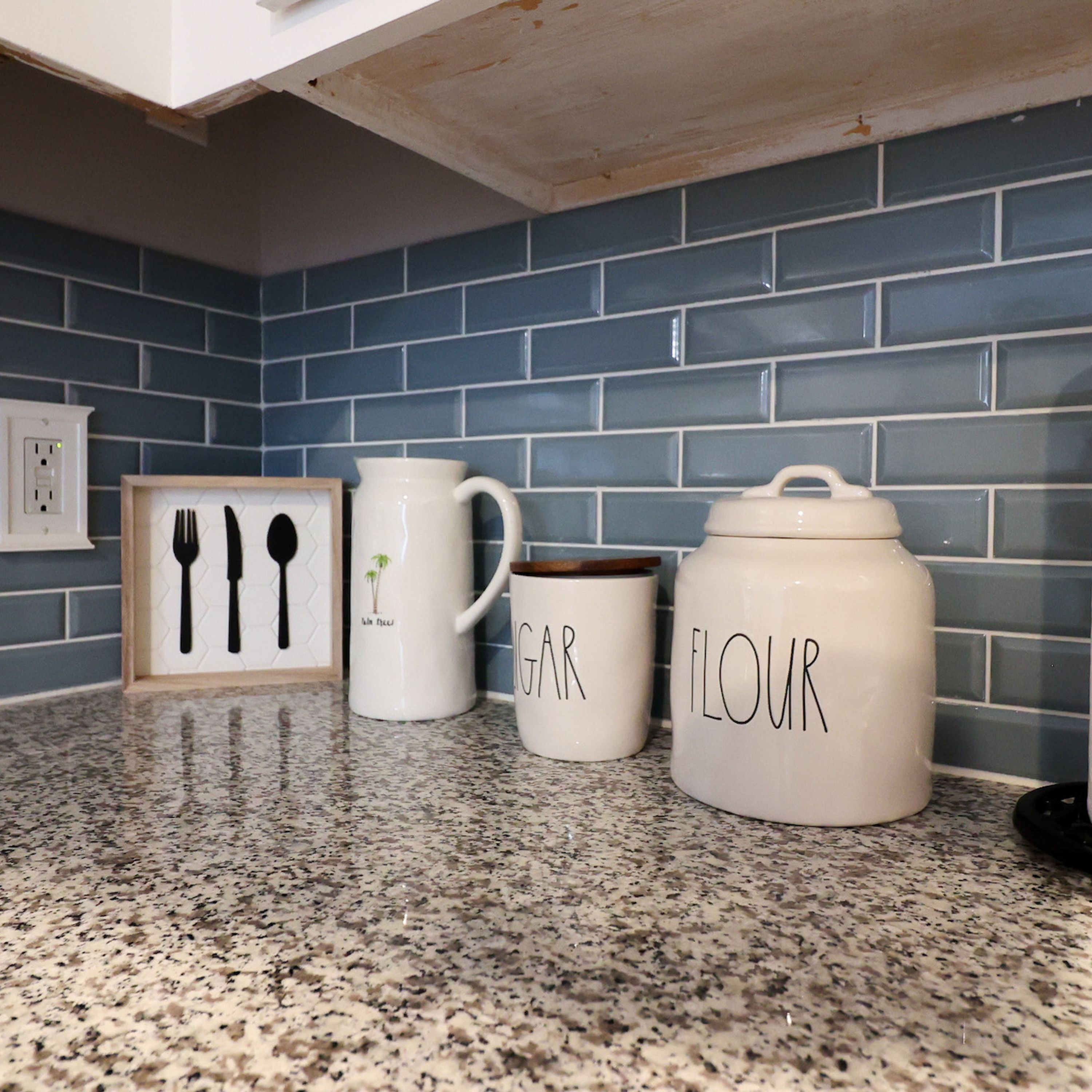 Eine Küchenarbeitsplatte mit einem gerahmten Bild von Besteck, einer Tasse mit Palmenmotiv und einem großen weißen Behälter mit der Aufschrift 'FLOUR'.