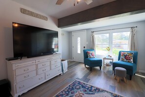 Un salon doté d’un téléviseur à écran plat, d’une commode blanche, de deux fauteuils bleus, d’un tapis à motifs et d’une grande fenêtre avec des rideaux.