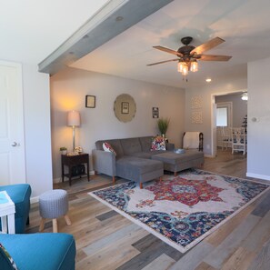 Un salon spacieux avec un grand tapis, un ventilateur de plafond et un coin repas à l’arrière-plan.