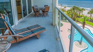 A balcony with lounge chairs and a table, overlooking a beach and a swimming pool. - Seas the Day in This Luxury Condo at Diamond Beach Ocean Views Incredible Pools Amenities (Galveston)