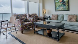 A spacious living room with a large window overlooking the ocean, a ceiling fan, and a painting of a toucan on the wall. - Seas the Day in This Luxury Condo at Diamond Beach Ocean Views Incredible Pools Amenities (Galveston)
