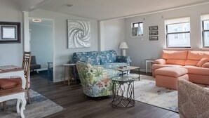 A spacious living room with a blue floral sofa, a peach-colored sofa, and a coffee table. There is a dining area with a wooden table and chairs. - Walk to the Beach Ocean Views Yard Game Room (Galveston)