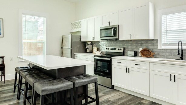 A modern kitchen with white cabinets, a stainless steel refrigerator, a microwave, an oven, and a coffee maker. There is a large island with bar stools and a ceiling fan. - Adventure Awaits Close to the Beach (Galveston)