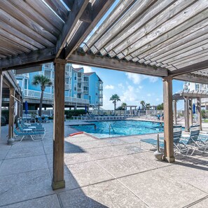 A pool area with a wooden pergola, lounge chairs, and a view of a blue building in the background. - Easy Beach Access Charming Beachside Condo (Galveston)