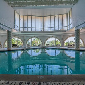 An indoor swimming pool with clear water, surrounded by lounge chairs and a high ceiling with large windows. - Western Waves Dreamy Diamond Beach Condo - Mind-blowing Views Amenities (Galveston)