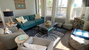 A spacious living room with a teal sofa, a grey sofa, and a round ottoman. There is a coffee table, a bookshelf, and a lamp. Large windows with curtains provide natural light. - Beachside Paradise Retreat With Luxury (Galveston)