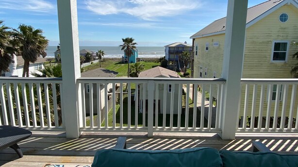A covered porch with a view of the ocean, a grassy yard, and a yellow house. - Beachside Paradise Retreat With Luxury (Galveston)