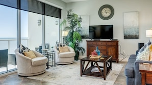 A spacious living room with a large window, a ceiling fan, a flat-screen TV, and a modern fireplace. - Welcome to Your Ultimate Getaway (Galveston)