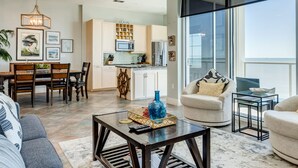 A spacious living room with a modern sofa, a coffee table, and a dining area with chairs and a table. There is a kitchen in the background with a microwave and cabinets. - Welcome to Your Ultimate Getaway (Galveston)