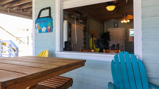A beach bar with a wooden table and chairs, a window with a view inside, and a sign above the window. - In-home Bar Lounge and Arcade Stay Entertained at The Sea Shed (Galveston)