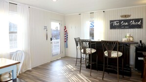 A dining area with a wooden table and chairs, a kitchen area with a bar, and a sign that reads "The Sea Shed EST. 2021". - In-home Bar Lounge and Arcade Stay Entertained at The Sea Shed (Galveston)