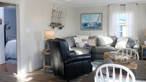 A modern living room with a black leather sofa, a grey couch, and a white dining table with a white chair. There is a ceiling fan, a painting of a sailboat, and a lamp on a side table. - In-home Bar Lounge and Arcade Stay Entertained at The Sea Shed (Galveston)