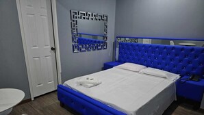 A bedroom with a blue upholstered bed, a white bedside table, a small round table, and a mirror with a geometric frame. - RESIDENCIAL LA MALVARROSA (Panama City)