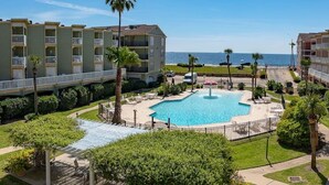 A resort with a central swimming pool, surrounded by palm trees and a view of the ocean. - Charming Waterfront Condo (Galveston)