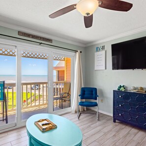A room with a ceiling fan, a TV, a blue chair, a wooden cabinet, and a view of the beach through glass doors. - Charming Waterfront Condo (Galveston)
