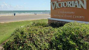 A sign for The Victorian Condominiums Meeting & Event Centers with a view of the ocean and beach in the background. - Charming Waterfront Condo (Galveston)